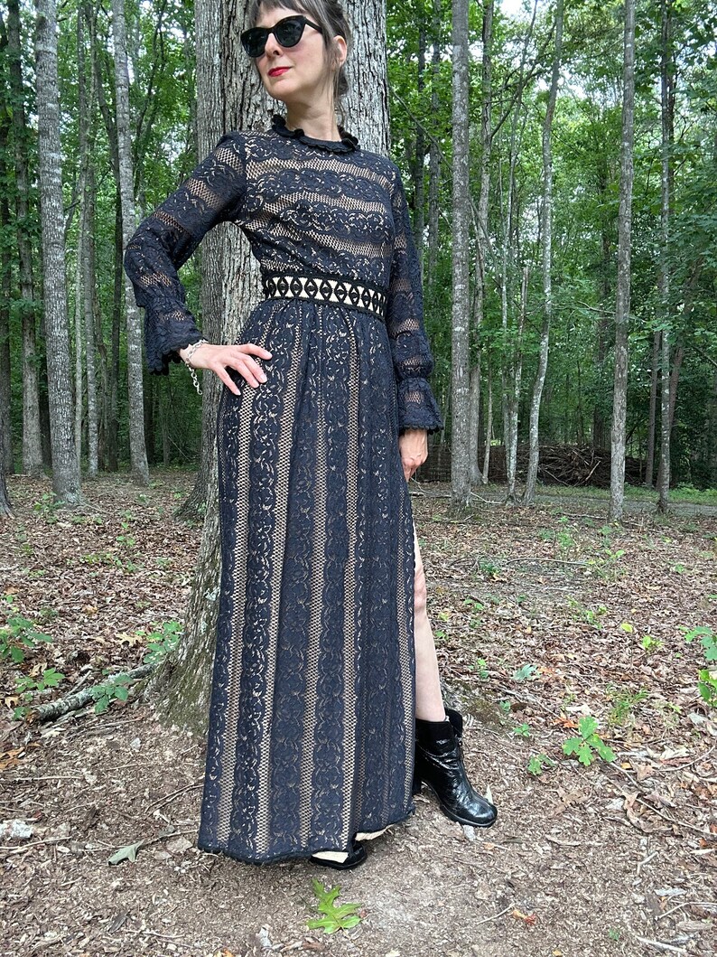May include: A black lace maxi dress with a high neck, long sleeves, and a fitted waist. The dress has a black belt with a geometric design and a thigh-high slit. The model is wearing black boots.