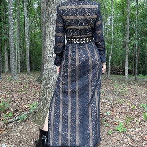 May include: A black lace maxi dress with long sleeves and a high neckline. The dress has a fitted bodice and a flowing skirt with a slit up the side. The dress is belted at the waist with a black belt.