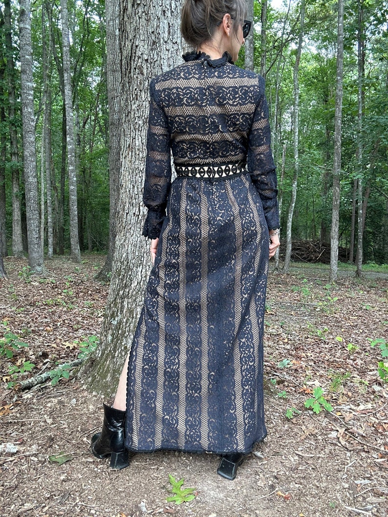 May include: A black lace maxi dress with long sleeves and a high neckline. The dress has a fitted bodice and a flowing skirt with a slit up the side. The dress is belted at the waist with a black belt.