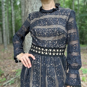 May include: A black lace dress with long sleeves and a high neckline. The dress has a fitted bodice and a flowing skirt. The lace is delicate and intricate, and the dress is accented with a black ribbon belt.