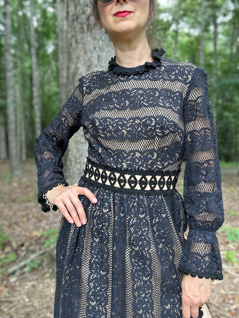 May include: A black lace dress with long sleeves and a high neckline. The dress has a fitted bodice and a flowing skirt. The lace is delicate and intricate, and the dress is accented with a black ribbon belt.