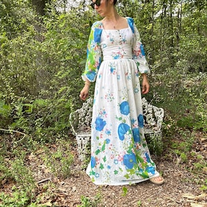 May include: A white maxi dress with a floral pattern featuring blue and pink flowers. The dress has long sleeves with a blue and green floral design, a square neckline, and a fitted bodice. The dress is worn outdoors.