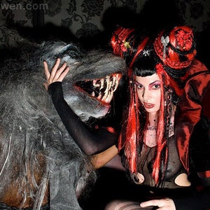 May include: A woman with red and black hair, wearing a red and black dress, poses with a wolf-like creature. The woman has dark makeup and is wearing a black choker necklace. The creature has a large, open mouth with sharp teeth.
