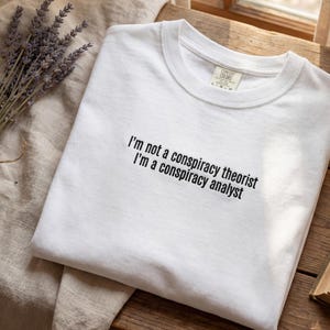 May include: White t-shirt with the text "I'm not a conspiracy theorist I'm a conspiracy analyst" in black. The shirt is folded on a light-coloured surface, with lavender and a book visible in the background.