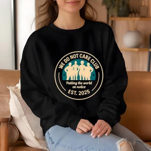 Puede incluir: Sudadera negra de cuello redondo con un gr&aacute;fico circular. El gr&aacute;fico presenta el texto "WE DO NOT CARE CLUB" alrededor de un dise&ntilde;o turquesa y beige con las palabras "Putting the world on notice EST. 2025".