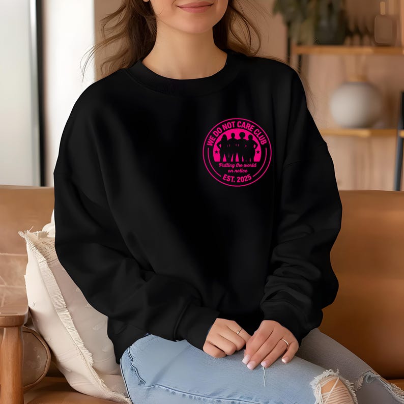 Puede incluir: Sudadera negra con un gr&aacute;fico circular rosa en el pecho. El gr&aacute;fico presenta el texto "WE DO NOT CARE CLUB" alrededor de una silueta de personas, con "Putting the world on notice" y "EST. 2025" debajo. La sudadera tiene cuello redondo.