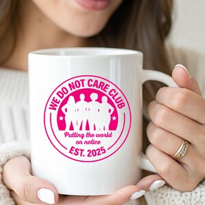 Puede incluir: Taza de cer&aacute;mica blanca con un gr&aacute;fico rosa que dice "WE DO NOT CARE CLUB" alrededor de una silueta de personas. Tambi&eacute;n incluye el texto "Putting the world on notice. EST. 2025".