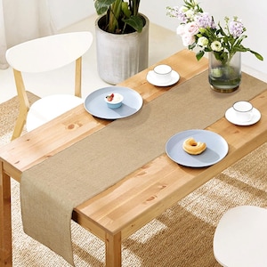 May include: A rectangular tan burlap table runner laid across a light wood dining table. The table is set with two light blue plates, two white teacups on saucers, a small bowl with berries, and a donut. A glass vase with flowers sits nearby.
