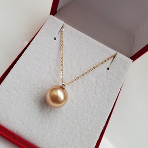 K&ouml;nnte beinhalten: Eine goldene Kettenkette mit einem gro&szlig;en goldenen Perlenanh&auml;nger. Die Perle ist mit einem kleinen Diamanten besetzt. Die Kette wird in einer roten Samtbox pr&auml;sentiert.