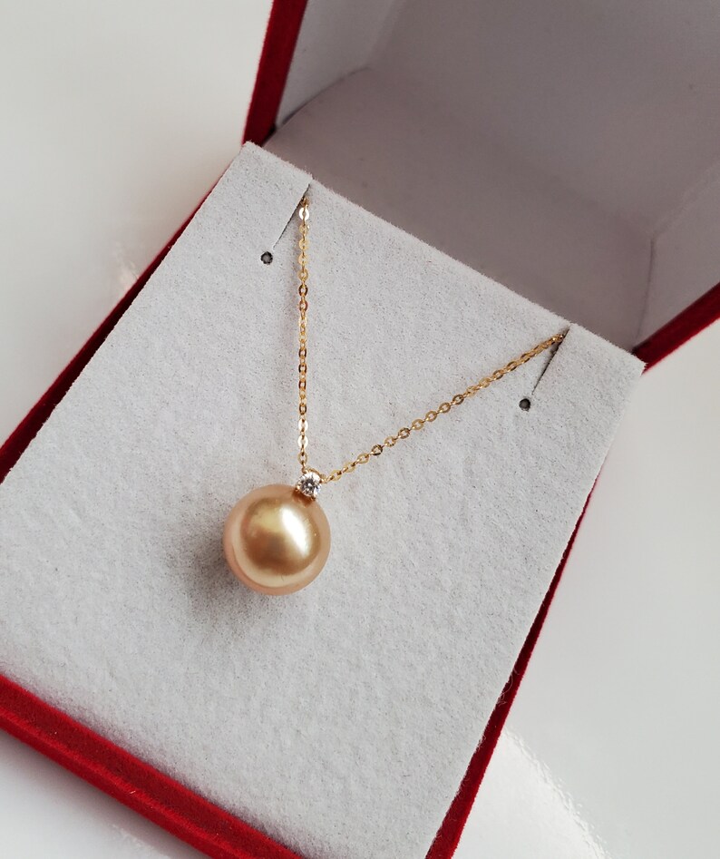 K&ouml;nnte beinhalten: Eine goldene Kettenkette mit einem gro&szlig;en goldenen Perlenanh&auml;nger. Die Perle ist mit einem kleinen Diamanten besetzt. Die Kette wird in einer roten Samtbox pr&auml;sentiert.
