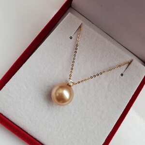 K&ouml;nnte beinhalten: Eine goldene Kettenkette mit einem gro&szlig;en goldenen Perlenanh&auml;nger. Die Perle befindet sich in einer roten Samt-Schmuckschatulle.