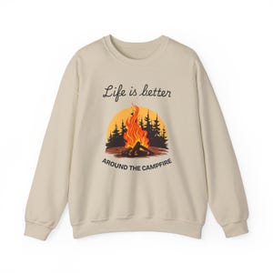 Könnte beinhalten: Ein beiger Pullover mit einem Lagerfeuer-Motiv und dem Text "Life is better around the campfire".