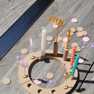 Könnte beinhalten: Ein Geburtstagskuchenring mit Kerzen und Konfetti. Der Ring besteht aus hellbraunem Material und hält eine weiße Kerze, eine rosa Kerze und zwei Spiral-Kerzen in Orange und Türkis. Goldkonfetti und das Wort "eins" sind ebenfalls vorhanden.