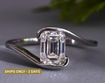 May include: A silver-toned bypass ring featuring a single emerald-cut clear gemstone held securely by the curved ends of the band. The metal band has a smooth, polished finish that wraps around the central stone in a continuous, flowing design.