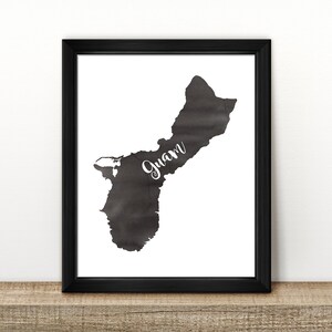 May include: A black and white print of the island of Guam in a black frame. The island is outlined in black and the name "Guam" is written in white cursive lettering.
