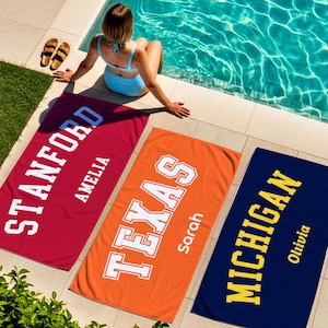 May include: Three personalized beach towels are displayed near a pool. One is red with "STANFORD" and "AMELIA" in white. The second is orange with "TEXAS" and "Sarah" in white. The third is navy blue with "MICHIGAN" and "Olivia" in yellow.