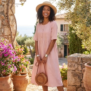 May include: A woman wearing a light pink dress with short sleeves and buttons down the front, holding a round woven handbag. She is also wearing a straw hat and silver bracelets. The background includes stone walls, potted flowers, and a building.
