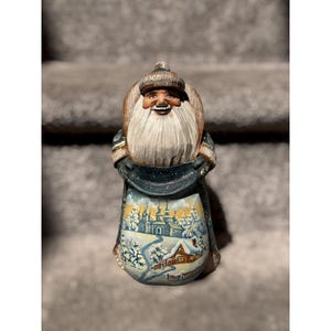 May include: A hand-carved wooden Santa Claus figurine. The Santa has a long white beard, a fur hat, and is holding a sack with a winter landscape scene. The scene depicts a church, houses, and snow-covered trees. The figurine is painted with various colors.