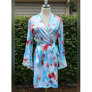 May include: A light blue satin robe with a floral pattern of red flowers. The robe has long sleeves with flared cuffs and a matching tie belt. The robe is displayed on a mannequin.