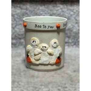 May include: A beige ceramic Halloween-themed container with three raised ghost figures and orange pumpkins. The words "Boo to you" are written on the rim. The container is cylindrical and has a slightly textured surface.