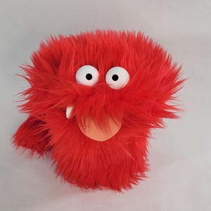 May include: A plush, red monster puppet with large white eyes, black pupils, and a small orange tongue. The puppet has a shaggy, furry texture and a single white tooth. The puppet is designed to be worn on the hand.