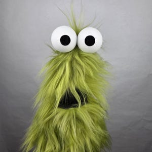 May include: A fuzzy, lime green puppet featuring long, shaggy synthetic fur, two large, round white eyes with black pupils, and a wide, dark, open-mouthed expression. The creature has a tall, upright structure with a textured, wild appearance.