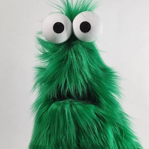 May include: A green, furry puppet with large, white eyes and black pupils. The puppet has a shaggy texture and a simple, open mouth. The puppet's fur is a vibrant, bright green color, and it has a whimsical, cartoonish appearance.