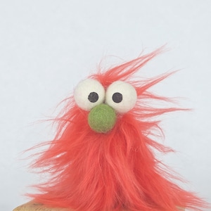 May include: A whimsical, handmade felt creature with a vibrant red, fluffy body and wild, spiky hair. It features large, white eyes with black pupils and a small, green nose. The creature has a playful, cartoonish appearance.