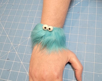 May include: A turquoise furry wrist cuff with two white and black googly eyes. The cuff is made of soft, plush material and is worn on the wrist. The background is a blue cutting mat.