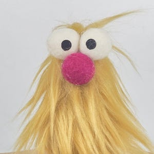 May include: A whimsical, handmade felt creature with large white eyes, black pupils, and a bright pink nose. The creature has long, yellow, shaggy hair. The design is playful and unique, perfect for a child's room or as a decorative item.