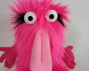 May include: A pink furry puppet with a long pink nose and large white eyes with black pupils. The puppet has black eyelashes and a pink mouth.