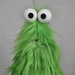 May include: A puppet featuring long, bright green synthetic fur covering its entire body. It has two large, round, white foam eyes positioned at the top, each with a solid black circular pupil in the center, creating a wide-eyed, expressive appearance.