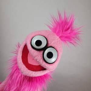 May include: A pink puppet with large, googly eyes and a wide, red-lined mouth. The puppet has a pink, fuzzy mohawk and matching fuzzy neck. The puppet's eyes are white with black pupils. The puppet is made of a soft, plush material.