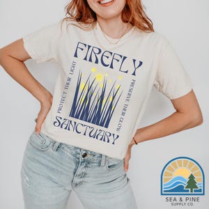 May include: A white t-shirt with a blue and yellow graphic design that says "FIREFLY PROTECT THEIR LIGHT PRESERVE THEIR GLOW SANCTUARY".