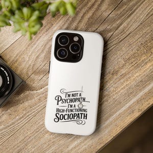 May include: White phone case with a black camera module and the text "I'm not a psychopath... I'm a high-functioning sociopath" in black script. The case is on a wooden surface.