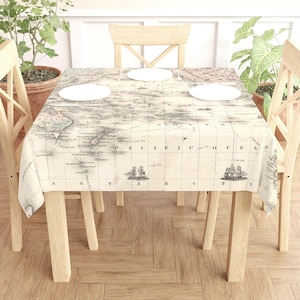 May include: A rectangular tablecloth featuring a vintage world map design in beige and gray tones. The map includes illustrations of sailing ships. The tablecloth is draped over a wooden table, with three white plates visible.