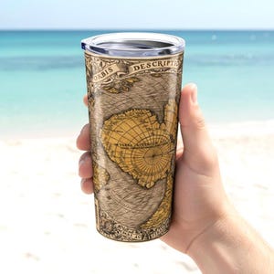 May include: A stainless steel travel tumbler featuring a vintage map design in shades of brown and gold. The tumbler has a clear lid and is held against a backdrop of a beach and ocean.