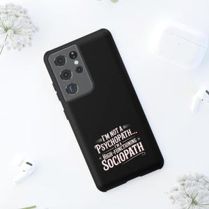 May include: Black phone case with the text "I'M NOT A PSYCHOPATH... I'M A HIGH-FUNCTIONING SOCIOPATH" in white. The case has a camera module on the upper left corner. The background is white with a pair of wireless earbuds and a charging case.