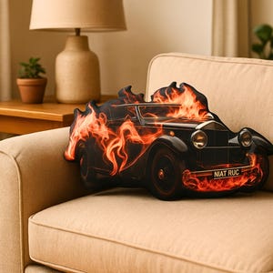 May include: A black vintage car-shaped pillow with flames engulfing the vehicle. The pillow has a license plate that reads "NIAT RUC". The pillow is resting on a beige sofa.