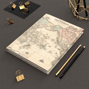 May include: A hardcover notebook with a vintage world map design. The notebook is open, displaying the map's details. Gold and black pencils, along with gold binder clips, are arranged around the notebook on a dark gray surface.