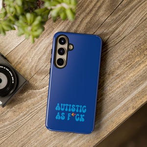 May include: A blue phone case with the text "AUTISTIC AS F*CK" in a retro font. The case is on a wooden surface, with a camera and plant in the background.