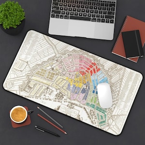 May include: A rectangular mousepad featuring a colorful vintage map of Amsterdam. The map is detailed with various colors and streets. A white computer mouse sits on the map, with a laptop, notebooks, and a cup of coffee nearby.