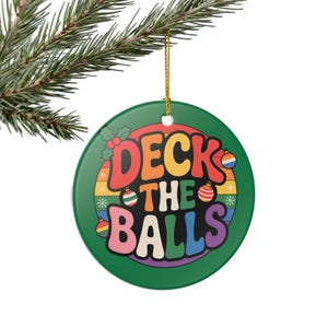 May include: A round, green Christmas ornament with the text "DECK THE BALLS" in rainbow colors. The ornament is decorated with snowflakes, holly, and rainbow-colored ornaments. A gold string is attached for hanging.