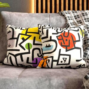 May include: Two decorative throw pillows with abstract designs. The pillows feature a white background with black lines and colorful shapes, including yellow, orange, green, and blue. They rest on a gray sofa.
