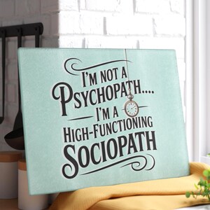 May include: A rectangular, light blue cutting board with the text "I'M NOT A PSYCHOPATH... I'M A HIGH-FUNCTIONING SOCIOPATH" in black script. A small pocket watch hangs from the top. The board rests on a yellow cloth.