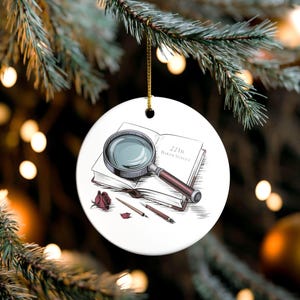 May include: White ceramic Christmas ornament with a Sherlock Holmes theme. The ornament features an open book with the text "221B Baker Street", a magnifying glass, a rose, and two pens. The ornament hangs from a gold string.