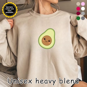 Könnte beinhalten: Beiges Unisex-Sweatshirt aus schwerem Mischgewebe mit einem lächelnden Avocado-Motiv. Die Avocado ist hellgrün mit einem braunen Kern und schwarzen Augen. Das Sweatshirt hat einen Rundhalsausschnitt und lange Ärmel. Der Text unten lautet "Unisex heavy blend". Mehrere Farben und Größen verfügbar.