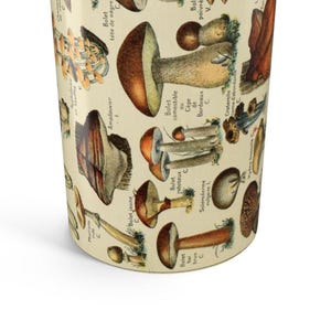 May include: A cylindrical tumbler with a beige background, covered in detailed illustrations of various mushrooms. The illustrations are in shades of brown, orange, and beige, with French text labels. The tumbler has a slightly angled perspective.