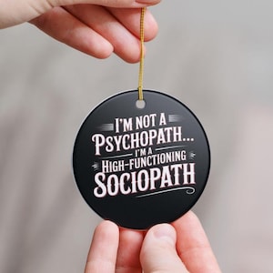 May include: A black round ornament with the text "I'M NOT A PSYCHOPATH... I'M A HIGH-FUNCTIONING SOCIOPATH" in white. The ornament is held by a gold string and is being held up by a person's hand.