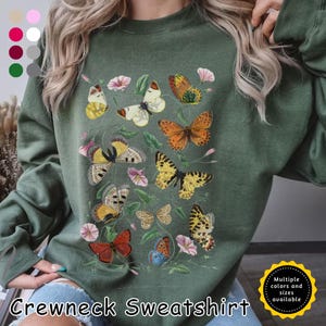 May include: Green crewneck sweatshirt featuring a butterfly and floral design. The design includes various colorful butterflies and pink flowers. The text "CREWNECK SWEATSHIRT" is at the bottom of the image. Multiple colors and sizes are available.